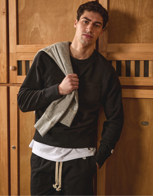 A photograph of a man looking at the camera while standing in front of a set of wooden lockers. He's wearing matching black sweatpants and a black crewneck sweatshirt over a white tee, with a gray sweatshirt tossed over his shoulder.