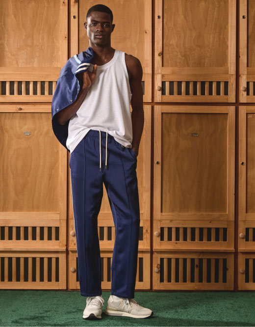 A photograph of a Black man looking at the camera while standing in front of a set of wooden lockers. He's wearing a white tank top, sneakers and blue drawstring pants, while holding a matching blue varsity jacket tossed over his shoulder.