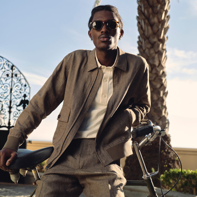 A photograph of a young man in sunglasses leaning against a bike outside, in front of a palm tree and a gate. The man is wearing glen plaid trousers, a glen plaid jacket, and a white sweater polo.