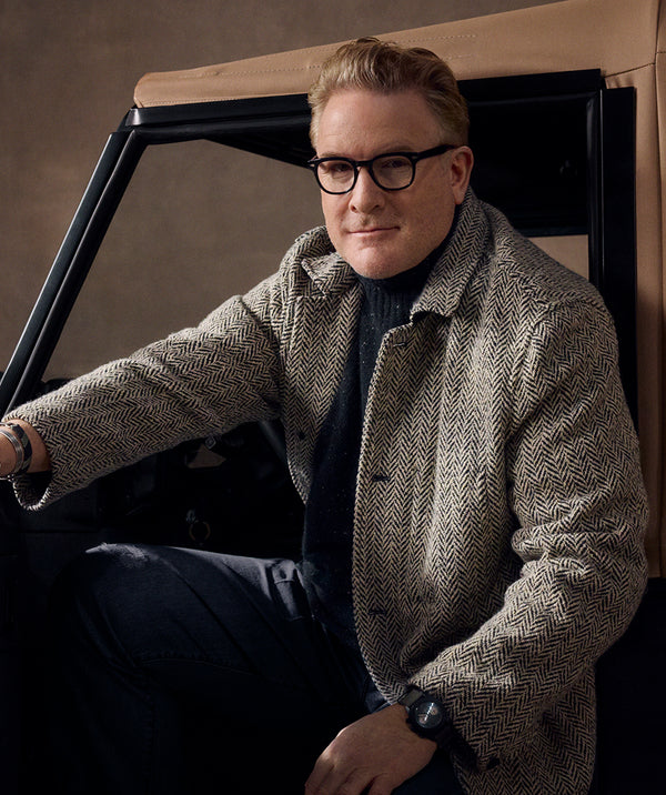 A close up of Todd Snyder sitting in the Balmoral Defender. He's wearing glasses, a herringbone tweed coat over a black turtleneck, dark trousers, and a wristwatch, photographed in dim, moody lighting.