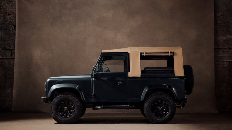 Side view of the Balmoral Defender soft-top in dark green, parked in a studio setting against a brown backdrop. Two-door pickup body style with a tan canvas convertible top rolled back and exposed roll bars, black wheel arch trim, black off-road wheels with all-terrain tires, and a rear-mounted spare tire. Shot in three-quarter profile from the side, low studio lighting.