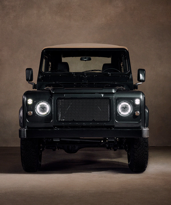 Front-on view of the Defender in the studio setting. Round LED headlights with illuminated halo rings, black mesh front grille, black bumper, black off-road wheels with all-terrain tires, side mirrors extended. Low studio lighting against a brown backdrop.