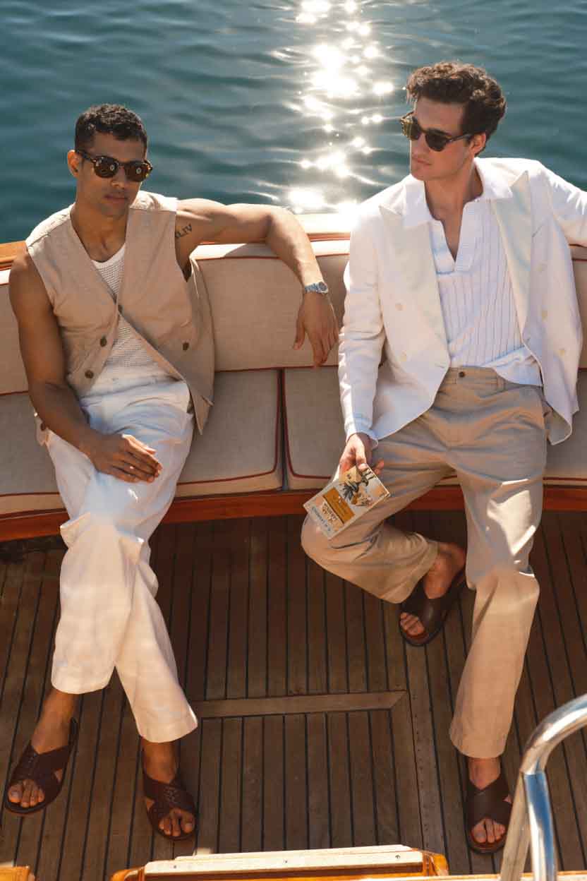 A photograph of two young men sit on a bench at the back of a yacht in the water. The one on the left is wearing sunglasses, a vest, and white chinos. The one on the right is wearing sunglasses, a white sweater polo, white suit jacket, and khaki chinos.