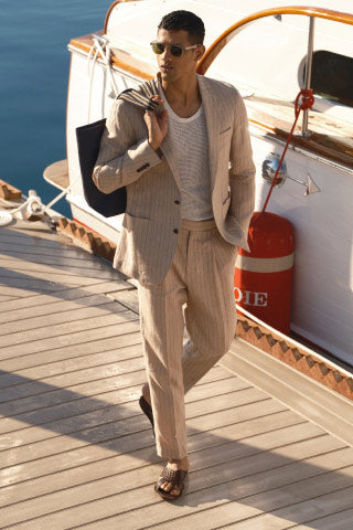A photograph of a man in a white mesh tank top, striped linen tan suit and sunglasses walking down the dock near a yacht and carrying a tote bag over his shoulder.