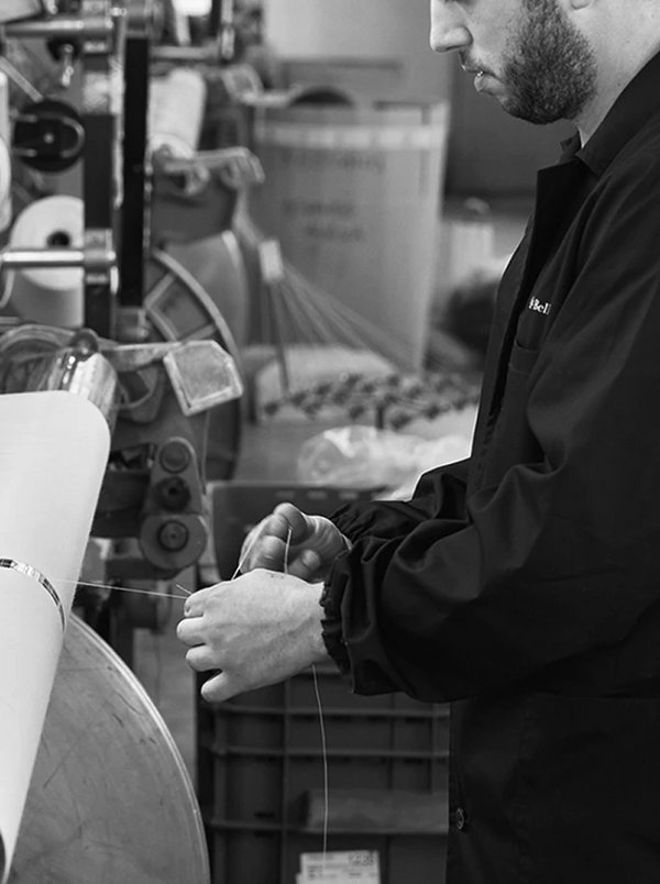 A bearded craftsman in a dark jacket threading or adjusting yarn on industrial textile equipment, photographed in black and white.