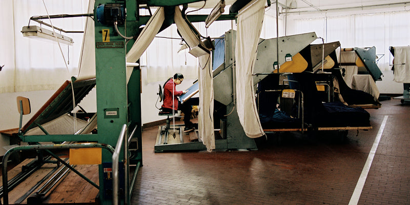 Large sheets of fabric are shown hanging in a clean mill, with a worker weaving fabric at a machine.