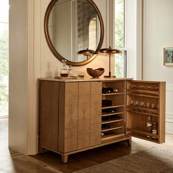 One door of the Tribeca marble and white oak bar cabinet is open revealing alcohol bottles and glasses. A top sits an Atelier aged brass lamp and Brandy. The Round Pied-a-Terre Gold wall mirror hangs above.